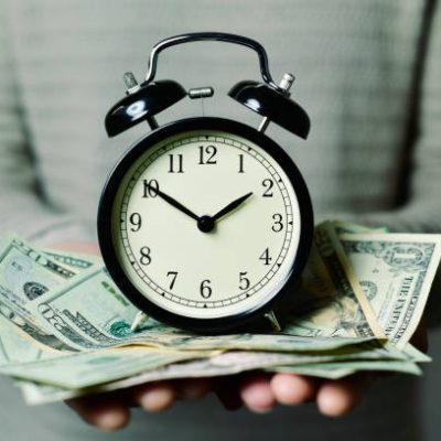 closeup of a young caucasian man with an alarm clock and many US dollar banknotes in his hands, depicting the idea that time is money